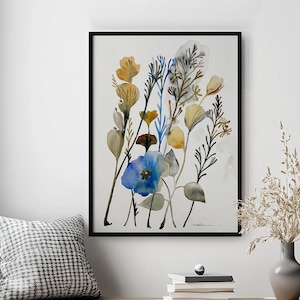 May include: A framed watercolour print of a botanical floral arrangement in blue, yellow, and green hues. The artwork hangs above a wooden bench with a cushion and woven baskets. A vase with dried plants is on the right.