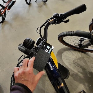 Pode incluir: Close-up de um guiador de bicicleta preto e azul com um visor digital e uma pega preta. A palavra "Nomad" é visível na bicicleta. O guiador é segurado por uma pessoa.