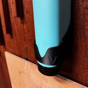 Op de afbeelding: Een turquoise waterfles wordt in een zwarte houder gehouden. De houder is op een houten oppervlak gemonteerd. De fles is cilindrisch en heeft een zwarte dop. De houder heeft een driehoekig ontwerp.