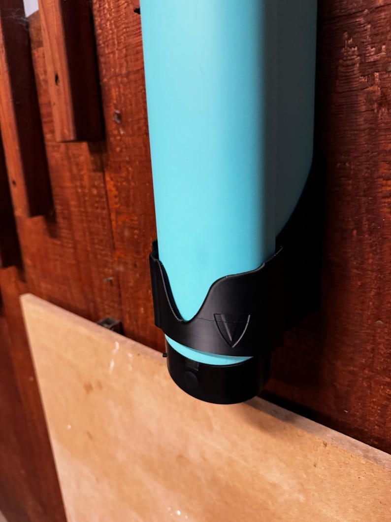 Op de afbeelding: Een turquoise waterfles wordt in een zwarte houder gehouden. De houder is op een houten oppervlak gemonteerd. De fles is cilindrisch en heeft een zwarte dop. De houder heeft een driehoekig ontwerp.
