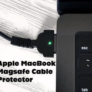 May include: Black Apple MacBook Magsafe cable protector plugged into a laptop. The protector has a green light and is connected to a black braided cable. The text "Apple MacBook Magsafe Cable Protector" is displayed in white.