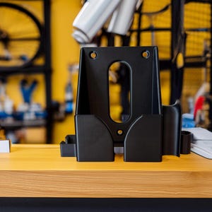 Pode incluir: Um suporte de repara&ccedil;&atilde;o de bicicletas preto, com formato retangular, apresentando uma abertura oval central e dois compartimentos laterais. O suporte &eacute; feito de material preto fosco e repousa sobre uma superf&iacute;cie de madeira. O fundo mostra pe&ccedil;as e ferramentas de bicicleta.