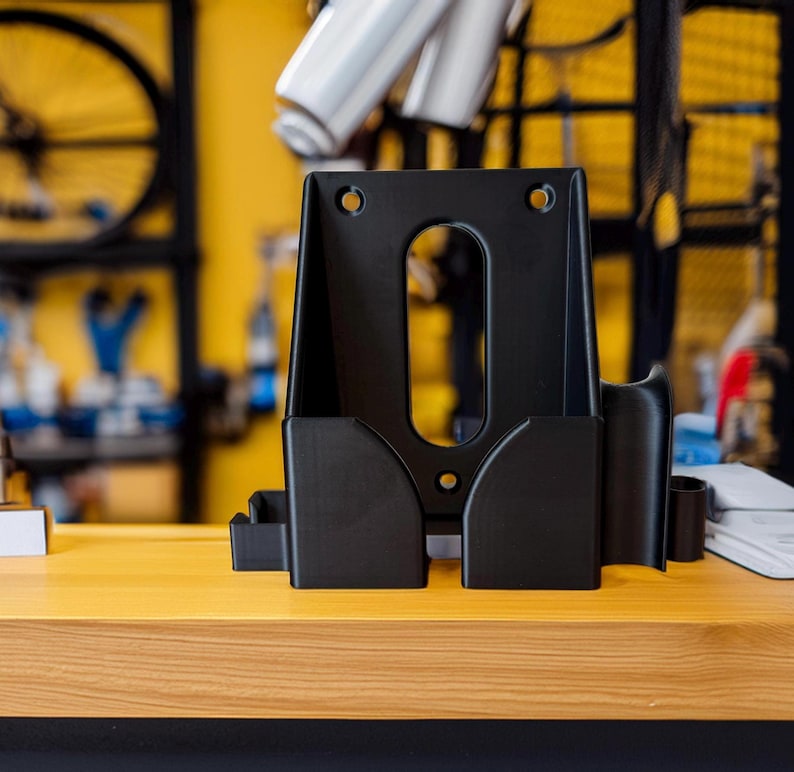 Pode incluir: Um suporte de repara&ccedil;&atilde;o de bicicletas preto, com formato retangular, apresentando uma abertura oval central e dois compartimentos laterais. O suporte &eacute; feito de material preto fosco e repousa sobre uma superf&iacute;cie de madeira. O fundo mostra pe&ccedil;as e ferramentas de bicicleta.
