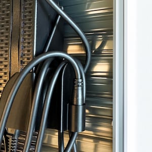 Pode incluir: Close-up de um cabo de alimenta&ccedil;&atilde;o e conector pretos, com um suporte de metal preto. O cabo &eacute; curvo e conectado a um dispositivo preto. O fundo &eacute; uma superf&iacute;cie met&aacute;lica ondulada.