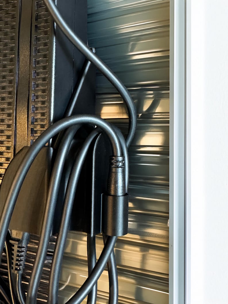 Pode incluir: Close-up de um cabo de alimenta&ccedil;&atilde;o e conector pretos, com um suporte de metal preto. O cabo &eacute; curvo e conectado a um dispositivo preto. O fundo &eacute; uma superf&iacute;cie met&aacute;lica ondulada.