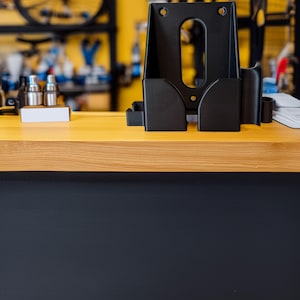 Pu&ograve; includere: Un supporto nero per la riparazione di biciclette con pi&ugrave; scomparti e fori di montaggio &egrave; posto su una superficie di legno chiaro. Il supporto &egrave; progettato per contenere utensili e accessori. Lo sfondo presenta una parete gialla e parti di biciclette.