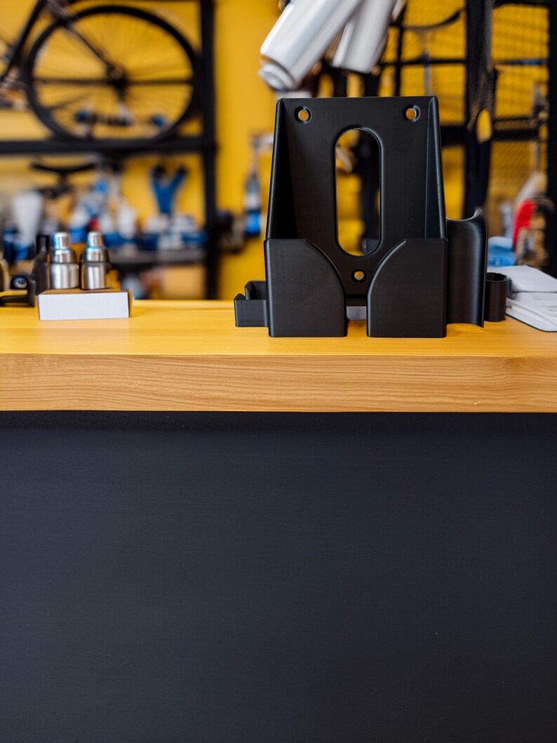 Pu&ograve; includere: Un supporto nero per la riparazione di biciclette con pi&ugrave; scomparti e fori di montaggio &egrave; posto su una superficie di legno chiaro. Il supporto &egrave; progettato per contenere utensili e accessori. Lo sfondo presenta una parete gialla e parti di biciclette.