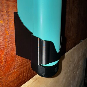 Op de afbeelding: Een turquoise waterfles in een zwarte wandhouder. De houder is gemaakt van een gebogen, solide materiaal. De fles is cilindrisch en lijkt van plastic te zijn. De houder is op een houten oppervlak gemonteerd.