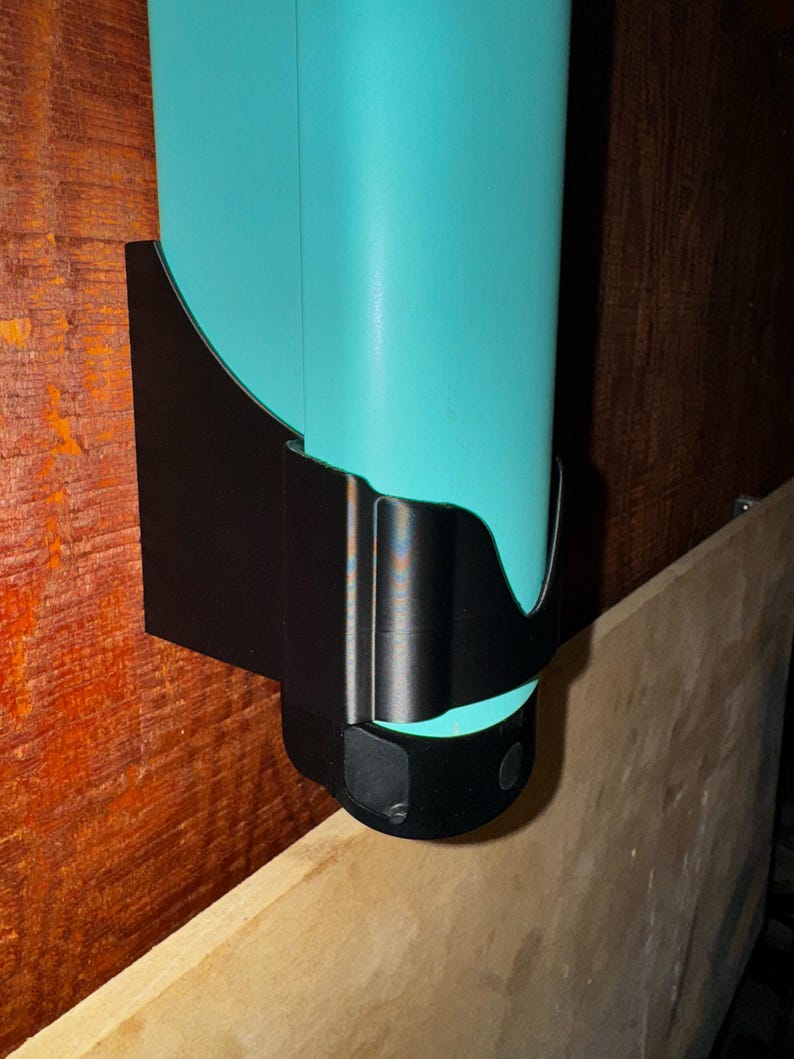 Op de afbeelding: Een turquoise waterfles in een zwarte wandhouder. De houder is gemaakt van een gebogen, solide materiaal. De fles is cilindrisch en lijkt van plastic te zijn. De houder is op een houten oppervlak gemonteerd.