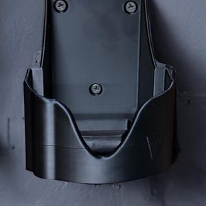 Op de afbeelding: Een donkergrijze, aan de muur gemonteerde stofzuigerhouder. De houder is gemaakt van plastic en heeft een gebogen voorkant met een sleuf voor de stofzuiger. Hij is met drie schroeven aan de muur bevestigd.