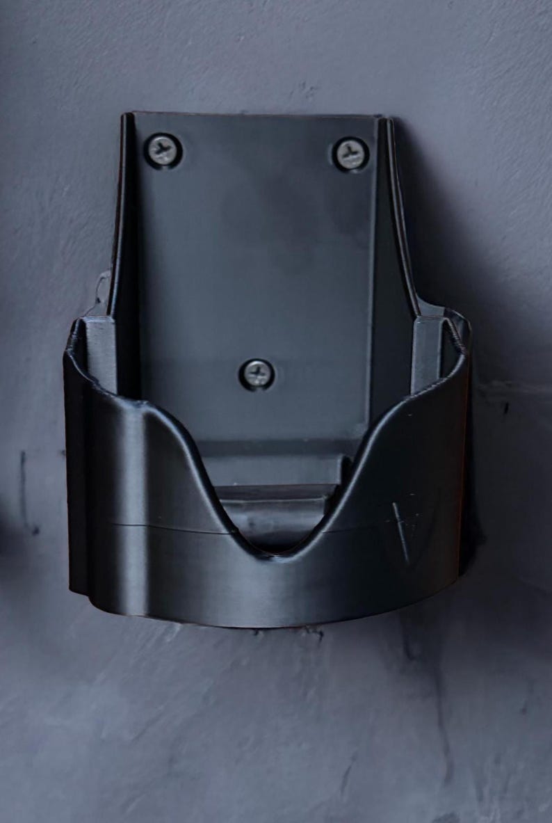 Op de afbeelding: Een donkergrijze, aan de muur gemonteerde stofzuigerhouder. De houder is gemaakt van plastic en heeft een gebogen voorkant met een sleuf voor de stofzuiger. Hij is met drie schroeven aan de muur bevestigd.