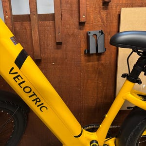 Pode incluir: Uma bicicleta el&eacute;trica amarela brilhante com a palavra "VELOTRIC" impressa na estrutura. A bicicleta est&aacute; montada numa parede de madeira, com um suporte preto para segurar a bicicleta. O assento &eacute; preto.