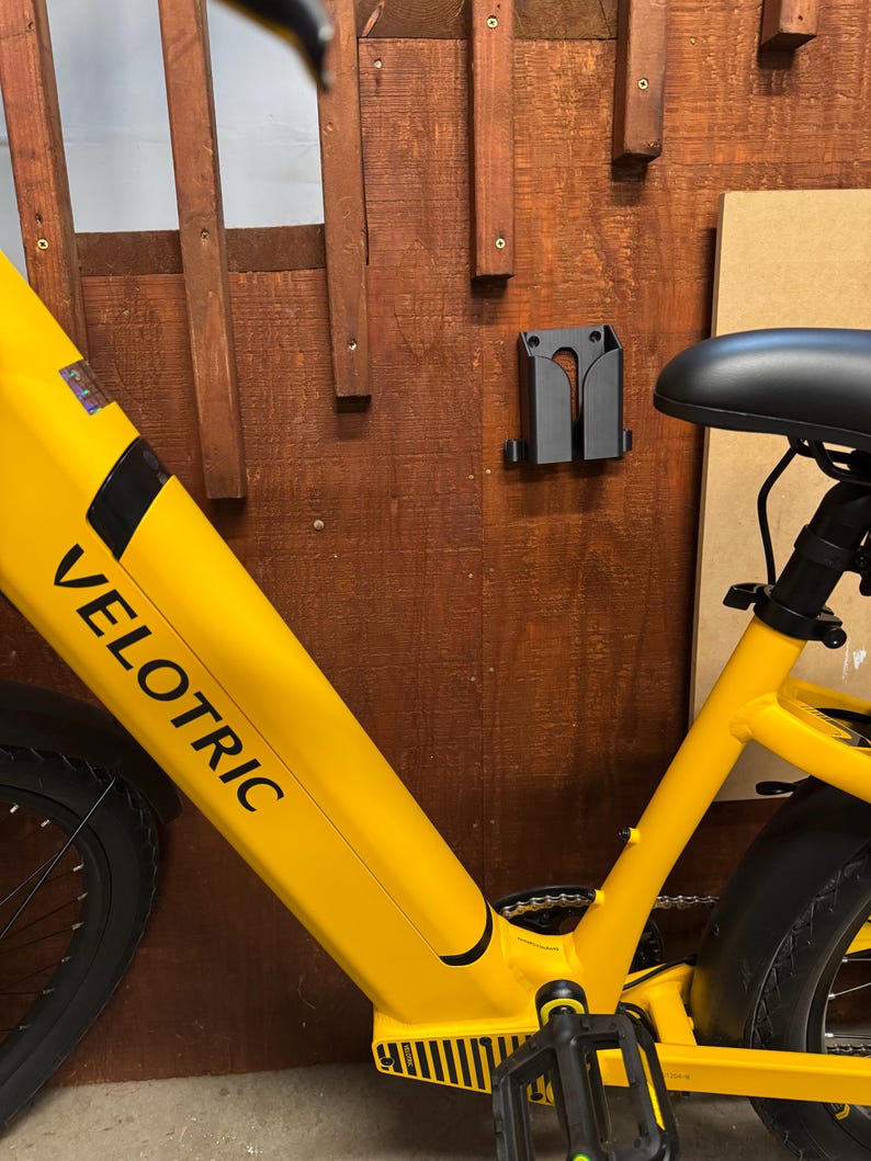 Pode incluir: Uma bicicleta el&eacute;trica amarela brilhante com a palavra "VELOTRIC" impressa na estrutura. A bicicleta est&aacute; montada numa parede de madeira, com um suporte preto para segurar a bicicleta. O assento &eacute; preto.