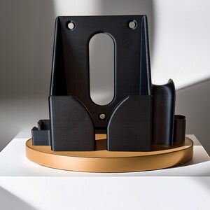Pode incluir: Um objeto geom&eacute;trico preto com um recorte oval e dois orif&iacute;cios na parte superior. Ele est&aacute; sobre uma base redonda e bronzeada. O objeto tem v&aacute;rios compartimentos e um design curvo, com uma est&eacute;tica minimalista.
