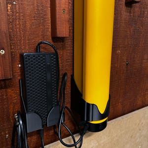 Op de afbeelding: Een gele laadstation met een zwarte basis en een zwarte voedingsadapter gemonteerd op een houten oppervlak. De voedingsadapter heeft een zwart snoer dat is aangesloten op een stopcontact.