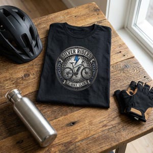 Puede incluir: Una camiseta negra con un gráfico circular que incluye el texto "SILVER RIDERS E-BIKE CLUB" rodeando la ilustración de una bicicleta eléctrica y un rayo azul. La camiseta se muestra junto a un casco negro, guantes y una botella.