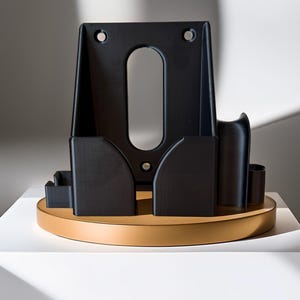 May include: A black, geometric object with an oval cutout and two holes at the top. It sits on a round, tan base. The object has several compartments and a curved design, with a minimalist aesthetic.