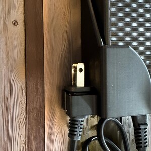 Pu&ograve; includere: Primo piano di una spina elettrica color oro inserita in un adattatore di alimentazione nero. L'adattatore &egrave; montato su una superficie di legno con assi verticali nei toni del marrone e del beige. I cavi elettrici neri sono collegati all'adattatore.