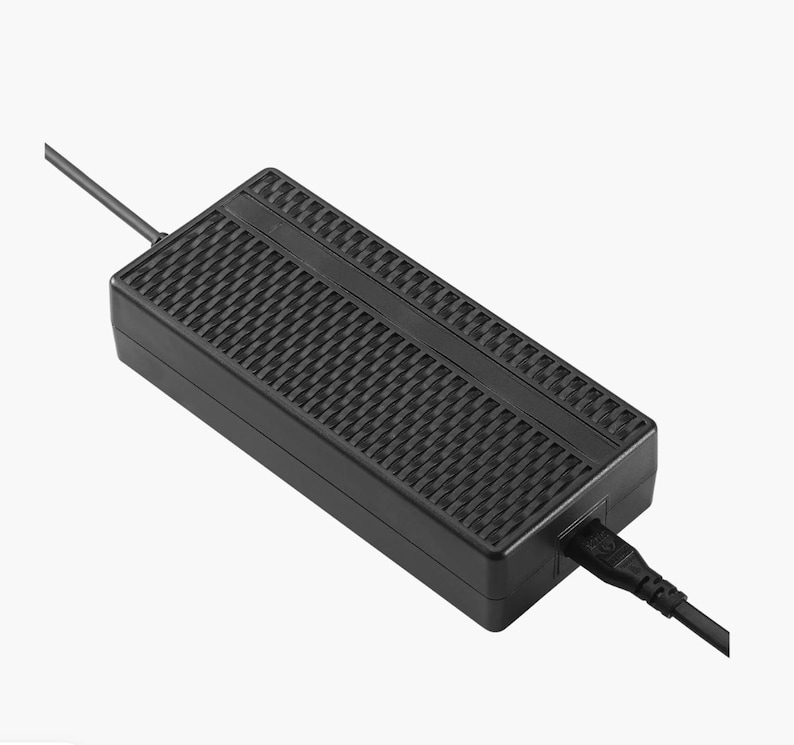 Pode incluir: Um adaptador de energia retangular preto com uma superf&iacute;cie texturizada e um cabo de alimenta&ccedil;&atilde;o que se estende de uma extremidade. O adaptador tem um padr&atilde;o de grade em sua superf&iacute;cie superior e uma porta para o cabo de alimenta&ccedil;&atilde;o.