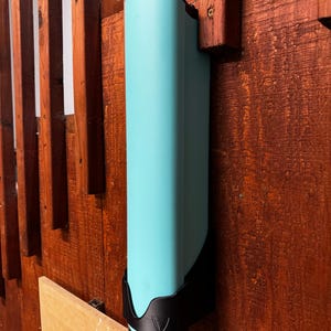 Op de afbeelding: Een turquoise, rechthoekige waterfles met een zwarte basis en bovenkant, gemonteerd op een houten oppervlak. De fles wordt op zijn plaats gehouden door een zwarte beugel. Het houten oppervlak heeft een roodbruine vlek.