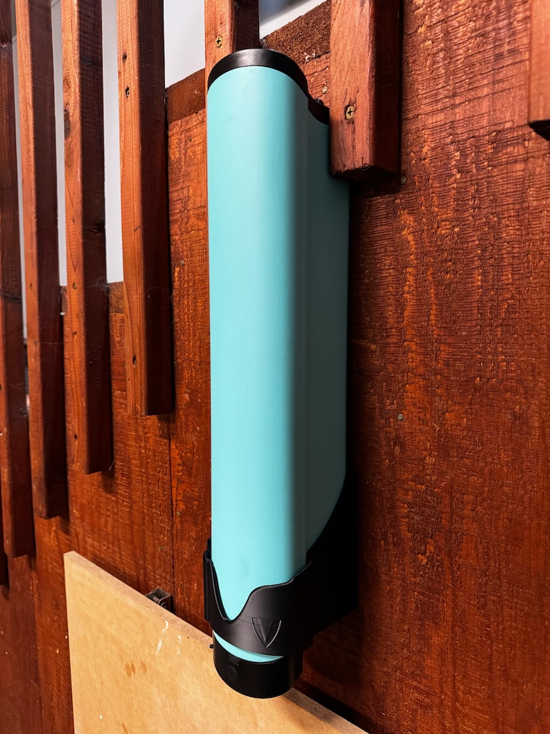 Op de afbeelding: Een turquoise, rechthoekige waterfles met een zwarte basis en bovenkant, gemonteerd op een houten oppervlak. De fles wordt op zijn plaats gehouden door een zwarte beugel. Het houten oppervlak heeft een roodbruine vlek.