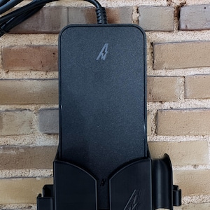 May include: A black charging device with a stylized "A" logo, mounted on a brick wall. The device is rectangular with a curved holder and a power cord extending from the top.
