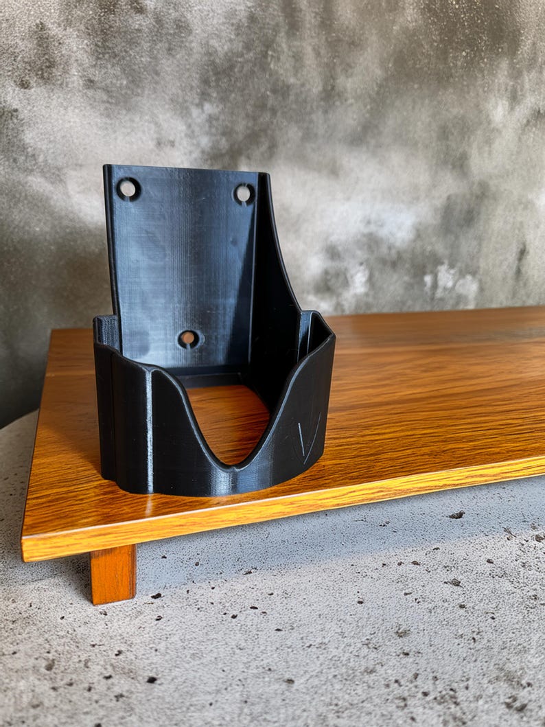 Op de afbeelding: Zwarte 3D-geprinte wandhouder voor een f&ouml;hn. De houder heeft een gebogen opening en twee montagegaten. Het staat op een houten oppervlak met een grijze achtergrond.