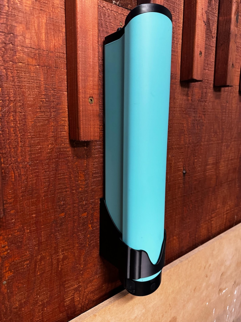 Op de afbeelding: Een turquoise, cilindrisch object gemonteerd op een houten oppervlak met een zwarte houder. Het object heeft een glad, gebogen ontwerp en wordt vastgezet door een zwarte beugel. Het houten oppervlak heeft een roodbruine tint.