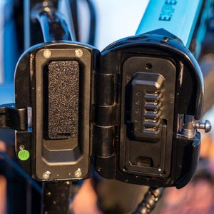 May include: A black plastic electric bicycle battery mount with an open hinged cover. The interior features a rectangular textured foam pad on the left side and a vertical row of five metal electrical contact pins on the right side.