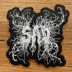 May include: Black patch with white text that reads "SAD" in a stylized font. The text is surrounded by a detailed, abstract design of white lines and shapes. The patch has a black border.