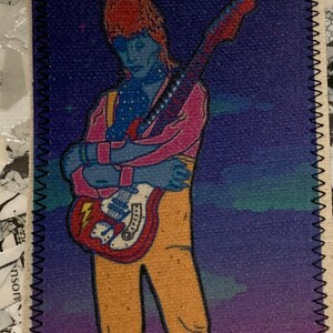 May include: A colorful patch featuring a stylized illustration of a musician with a guitar. The figure is depicted with blue skin, orange hair, and wearing a pink jacket and yellow pants. The guitar is red and white, set against a purple and blue background.