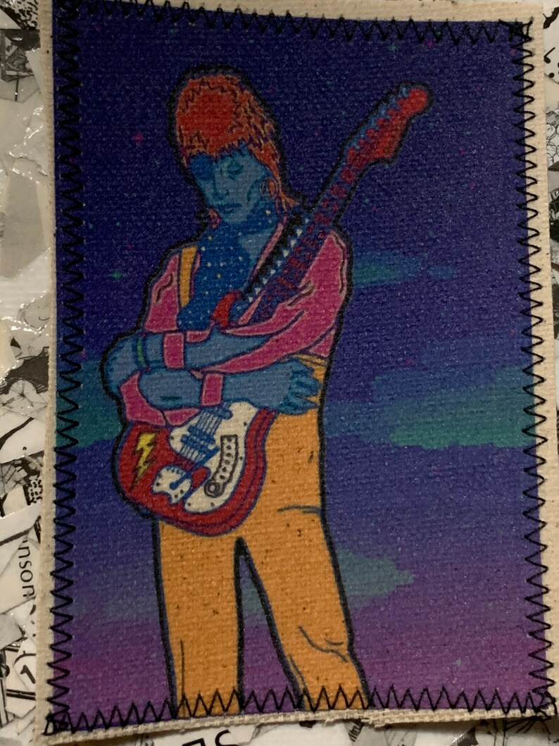 May include: A colorful patch featuring a stylized illustration of a musician with a guitar. The figure is depicted with blue skin, orange hair, and wearing a pink jacket and yellow pants. The guitar is red and white, set against a purple and blue background.