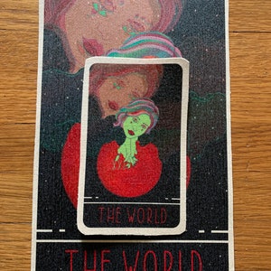 May include: A rectangular tarot card mat and a smaller card featuring a colorful illustration of a figure with green skin and red hair. The words "THE WORLD" are printed on both the mat and the card in red.
