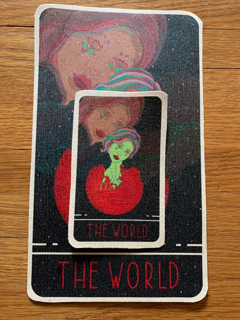 May include: A rectangular tarot card mat and a smaller card featuring a colorful illustration of a figure with green skin and red hair. The words "THE WORLD" are printed on both the mat and the card in red.
