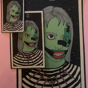 May include: Three tarot card-style patches featuring a green-skinned figure with a stitched mouth and a necklace of swords. The figure wears a black and white striped shirt. The largest patch has the Roman numeral VII in the upper corner.