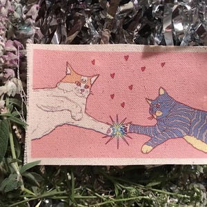 Op de afbeelding: Een rechthoekige stoffen patch met een kattenillustratie. Twee katten raken elkaar aan met hun poten, omringd door kleine rode hartjes op een roze achtergrond. E&eacute;n kat is wit met oranje accenten, de andere is blauw met strepen. De patch is omgeven door groen en glanzend zilver materiaal.