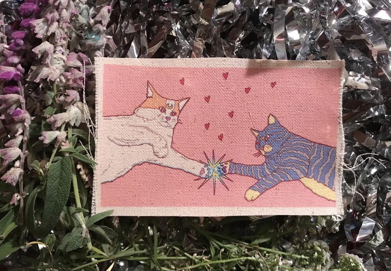 Op de afbeelding: Een rechthoekige stoffen patch met een kattenillustratie. Twee katten raken elkaar aan met hun poten, omringd door kleine rode hartjes op een roze achtergrond. E&eacute;n kat is wit met oranje accenten, de andere is blauw met strepen. De patch is omgeven door groen en glanzend zilver materiaal.