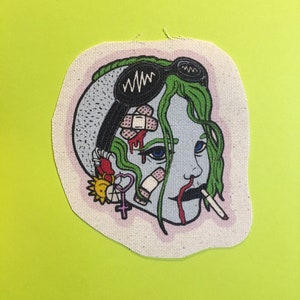 May include: A colorful embroidered patch featuring a cartoon illustration of a person's head with green hair, blue eyes, and a cigarette in their mouth. The head is covered in bandages, hearts, and other symbols.