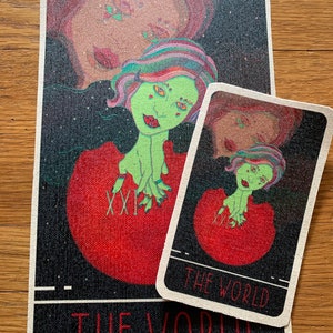 May include: Two tarot cards featuring a green-skinned figure with colorful hair emerging from a red orb. The larger card has the Roman numeral XXI and the words "THE WORLD" at the bottom. The smaller card has the same design and text.