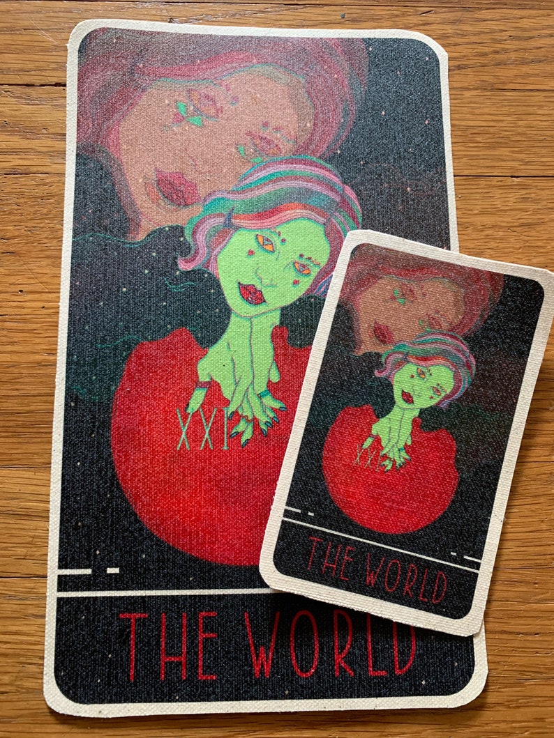 May include: Two tarot cards featuring a green-skinned figure with colorful hair emerging from a red orb. The larger card has the Roman numeral XXI and the words "THE WORLD" at the bottom. The smaller card has the same design and text.