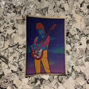 Può includere: Una toppa rettangolare in tessuto raffigurante un musicista con capelli arancioni e una camicia rosa, che tiene una chitarra rossa. Lo sfondo è un gradiente di blu e viola. La toppa è bordata da cuciture nere.