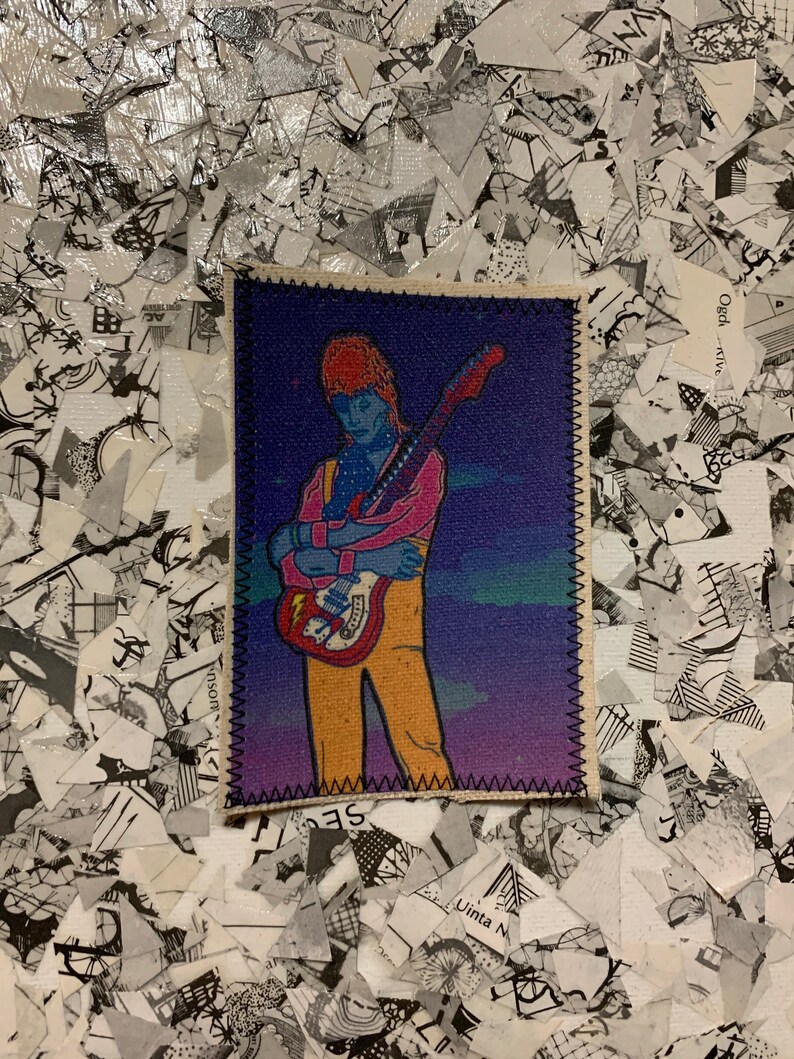 May include: A rectangular fabric patch featuring a stylized illustration of a musician with orange hair and a pink shirt, holding a red guitar. The background is a gradient of blue and purple. The patch is bordered with black stitching.
