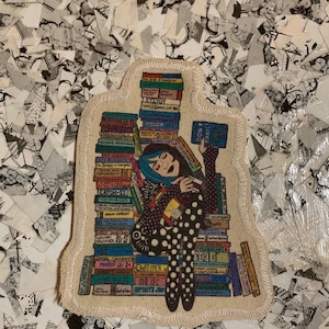 Peut inclure: Un patch blanc avec une illustration colorée d'une personne assise sur une pile de livres. La personne porte une robe à pois noirs et blancs et tient un livre qui dit "Ne paniquez pas".