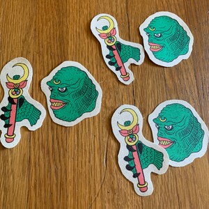 以下が含まれることがあります： 緑色のモンスターとピンクと黄色の杖が描かれたアイロン接着パッチが5枚。モンスターは緑色の体で、ピンクの口と黄色の目があります。杖はピンクの柄で、先端に黄色の星が付いています。