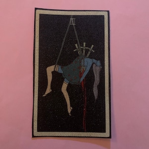 Può includere: Una toppa rettangolare con sfondo nero e bordo bianco. La toppa presenta un'illustrazione di una figura sospesa da corde, con spade che perforano la schiena. La figura indossa un vestito blu e ha lunghi capelli grigi. Linee rosse indicano sangue.