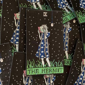 May include: Black rectangular patch featuring a tarot card illustration. The design depicts a figure in a blue dress reading a book under a starry sky. The text "THE HERMIT" is at the bottom, with the Roman numeral "IX" at the top.