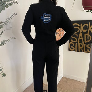May include: Black jumpsuit with a high collar and long sleeves. A blue and white embroidered design of a smiling coffee cup is on the back. The jumpsuit has a straight leg and is worn with black and white striped shoes.