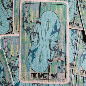 Könnte beinhalten: Die Tarotkarte Der Gehängte zeigt eine blaue Figur mit drei Augen, die kopfüber von einem Ast hängt. Die Karte ist von einem bunten Hintergrund mit einem Blumenmuster umgeben. Der Text "Der Gehängte" ist in Schwarz am unteren Rand der Karte gedruckt.