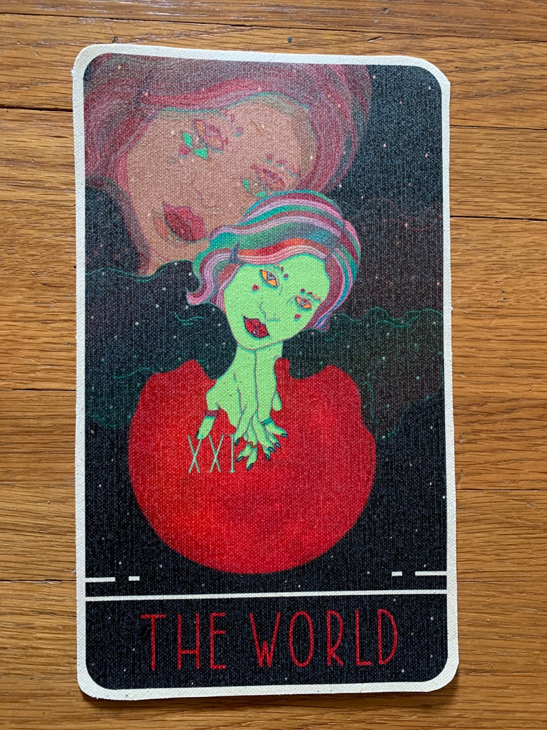 May include: A tarot card featuring a green-skinned figure with colorful hair emerging from a red orb. The card has a black background with stars and the text "THE WORLD" at the bottom. The Roman numeral "XXI" is visible.