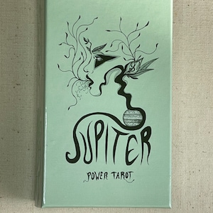 Puede incluir: Una caja de cartas rectangular de color verde menta claro con una ilustración de líneas negras de un perfil facial con ramas fluidas y un planeta. El texto JUPITER está impreso en letras negras grandes con POWER TAROT debajo.